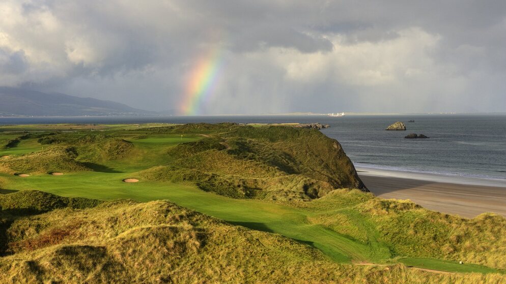 Coastal golf course in Ireland with a rainbow over the sea and rolling fairways.