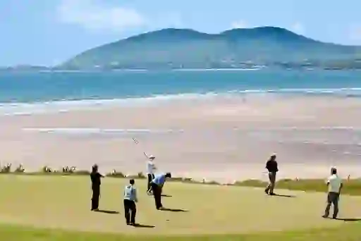 Golfers play by the shore at Waterville Golf Links with the Atlantic Ocean and mountains in the distance.