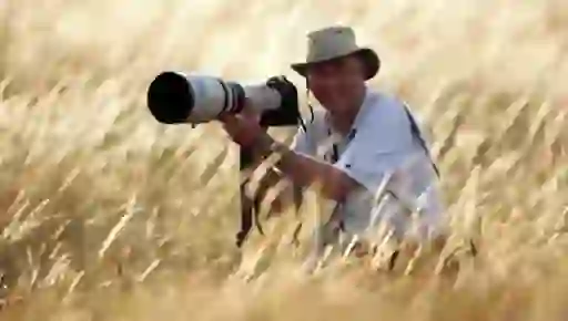 Golf photographer David Cannon kneels in tall grass with a long lens camera, smiling as he takes aim.