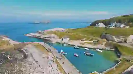 Ballintoy Harbour with small boats moored in clear blue water, stone paths, grassy cliffs and white cottages nearby.