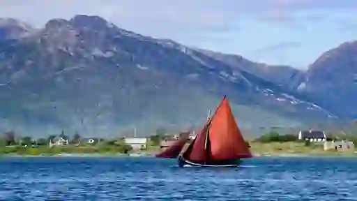 Traditional Galway Hooker boat with red sails sailing past cottages and Connemara mountains under blue sky.