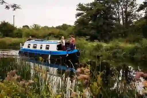 A blue canal boat with four people cruising along the Grand Canal in Sallins, County Kildare, surrounded by reeds and trees.