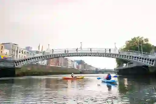 Two people kayaking the River Liffey under the Ha’penny Bridge with Dublin city buildings in the background.