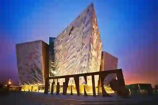 Titanic Belfast museum’s angular silver facade and large TITANIC sign illuminated at dusk in Belfast’s Titanic Quarter.