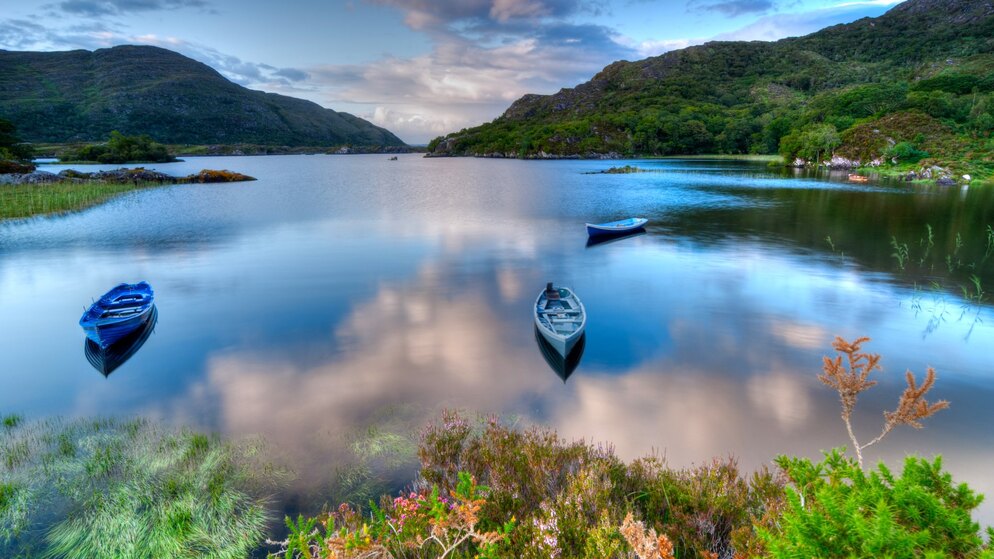 Calm lake in Killarney with wooded hills reflecting in water and small boats moored along grassy shore.