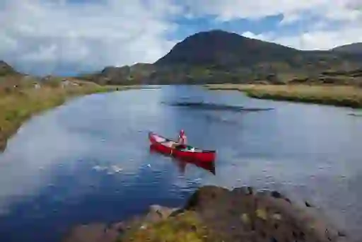 Solo paddler in a red canoe on a tranquil lake surrounded by rugged mountains in Killarney National Park.
