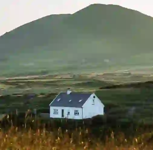 White cottage in the Ring of Kerry set against green fields and a softly lit mountain backdrop at sunset.