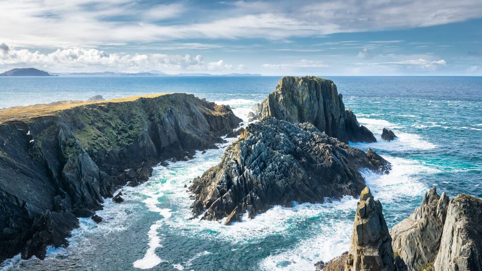 Rugged cliffs along the Wild Atlantic Way with waves crashing below, under a bright blue sky with scattered clouds.