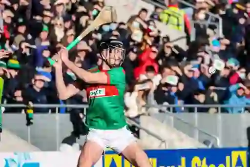 Hurler in green and red jersey striking a sliotar with a wooden stick during a match in Ireland.