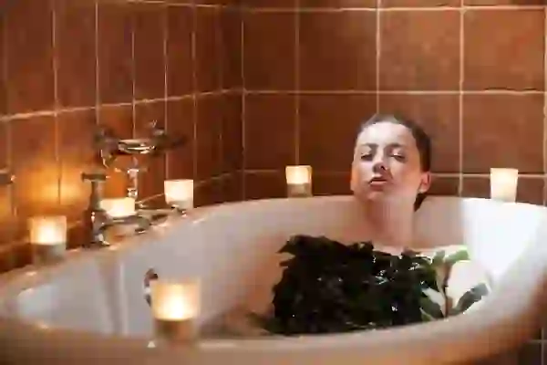 Person reclining in bath with seaweed on chest, eyes closed, surrounded by lit candles on ledge.