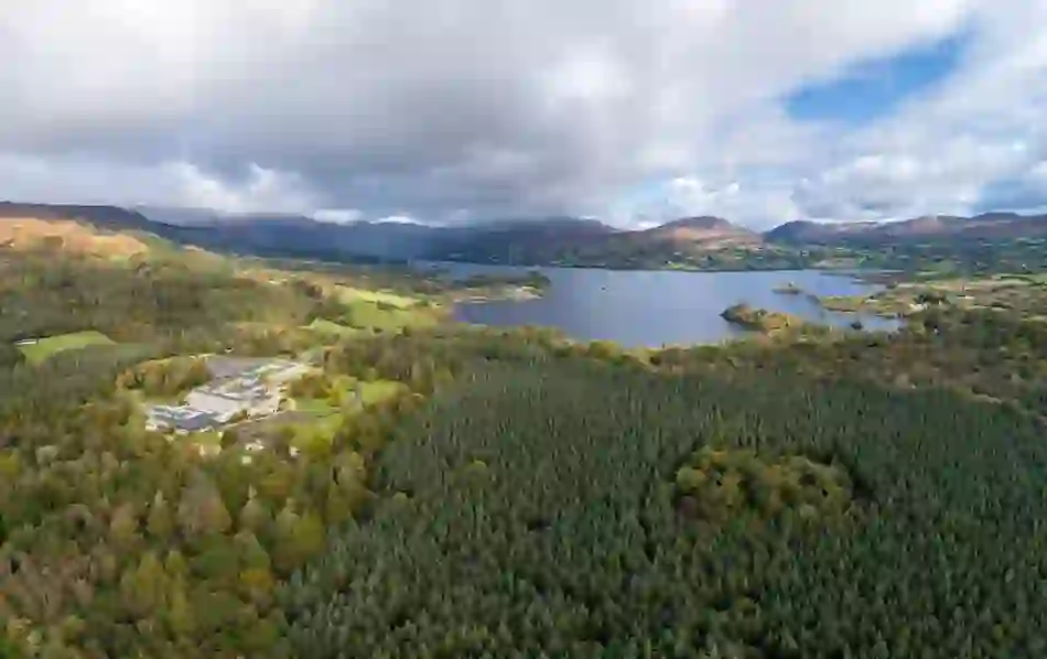 Drone view of Lough Eske Castle Hotel among forests beside lake and hills under partly cloudy sky.