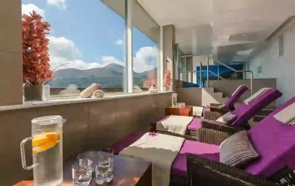 Relaxation room with purple loungers, rolled towels and mountain views through wide window.