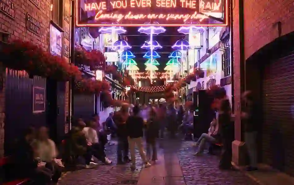 Cathedral Quarter alley in Belfast lit by neon umbrellas and signs, bustling with evening crowds.
