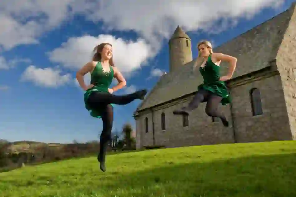 Two female Irish dancers in green dresses perform outdoors beside Saul Church in County Down.