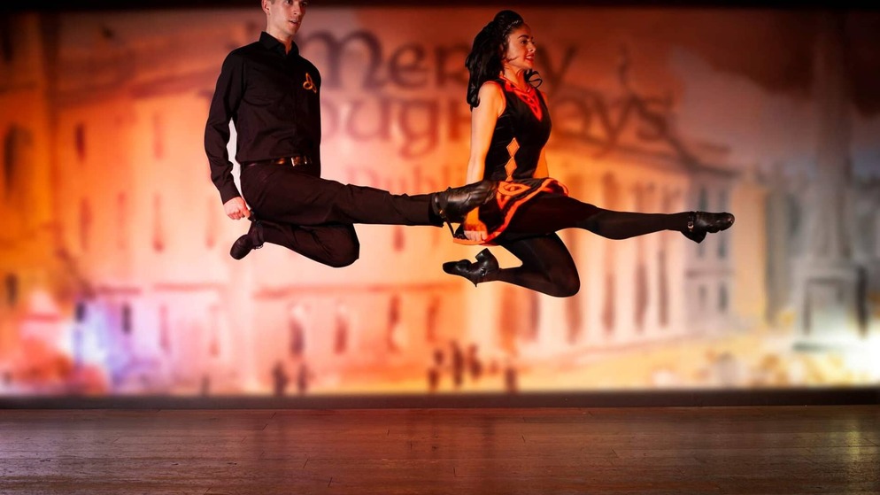 Irish dancers in black and red costumes leap mid-air during a traditional dance performance.