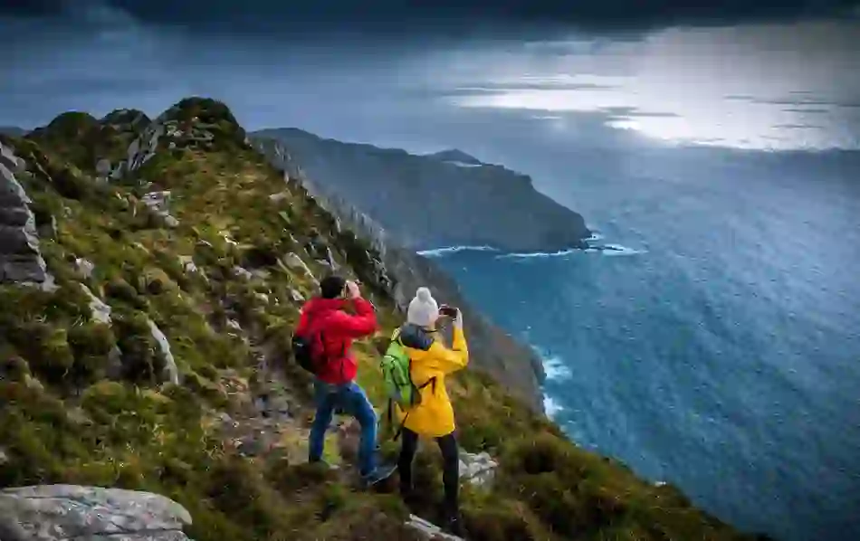 Hikers in bright jackets overlook dramatic Atlantic cliffs at Slieve League, County Donegal, under stormy skies.