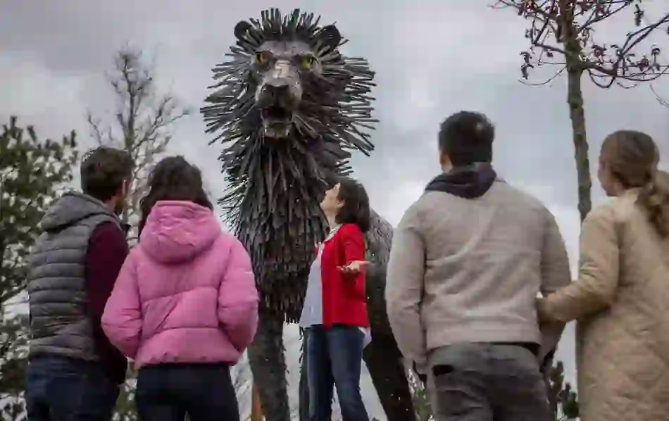 Visitors admire a towering sculpture of Aslan the lion at CS Lewis Square in Belfast, under a dramatic overcast sky.