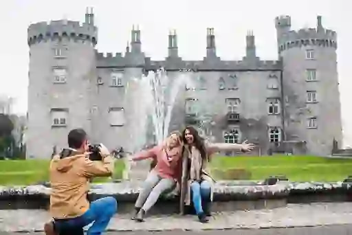 Tourists posing by the fountain in front of Kilkenny Castle on a cloudy day.