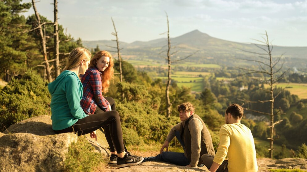 A group of young people sit chatting outdoors with a green valley backdrop.