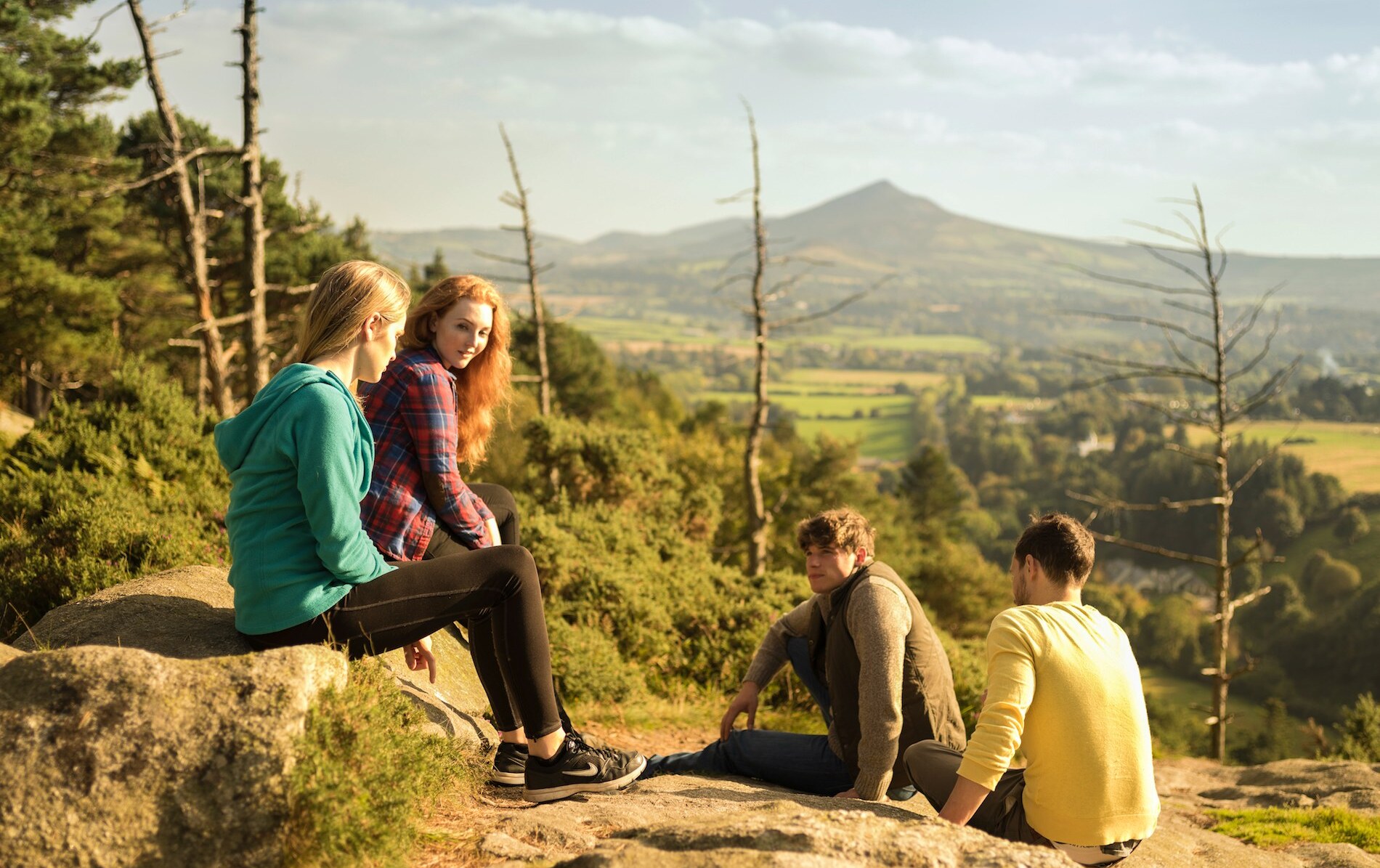 A group of young people sit chatting outdoors with a green valley backdrop.