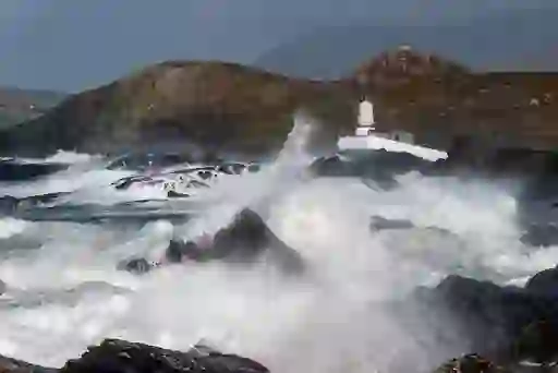 Waves crash against rocks near Valentia Island lighthouse, with hills in the background.