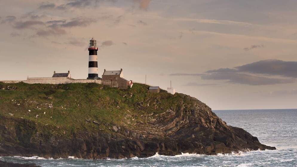Black-and-white striped lighthouse on rocky headland at sunset, with cottages and sea below.
