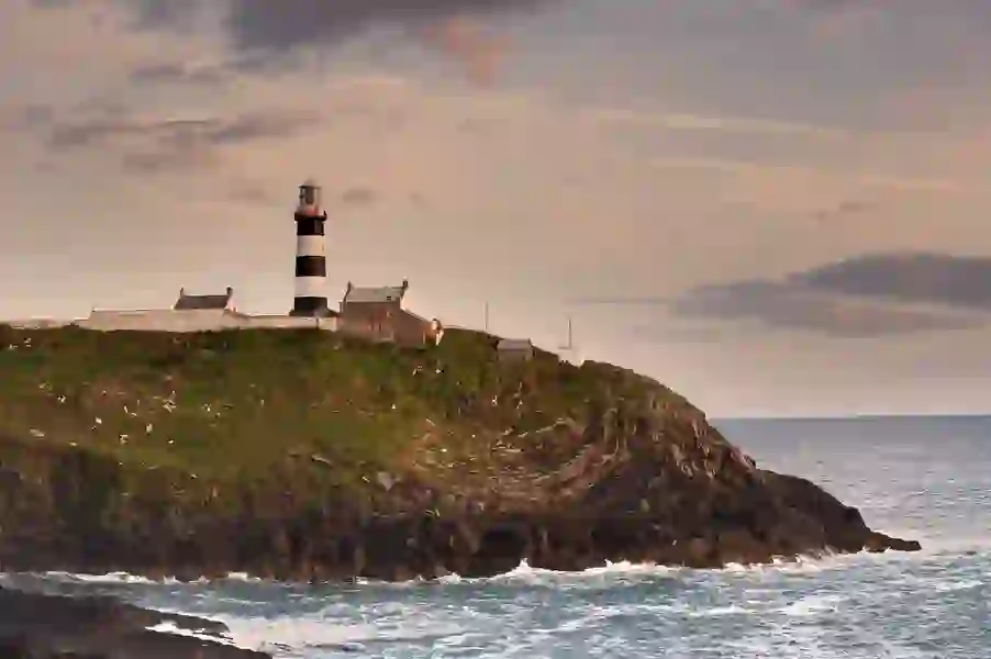 Black-and-white striped lighthouse on rocky headland at sunset, with cottages and sea below.