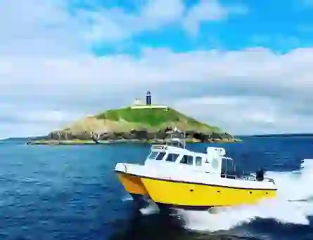 Yellow boat speeds past Ballycotton Island lighthouse, set on a grassy hill above the sea.