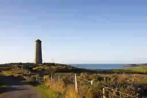 Tall stone Wicklow Head lighthouse stands by the coast on a clear day with blue sky.