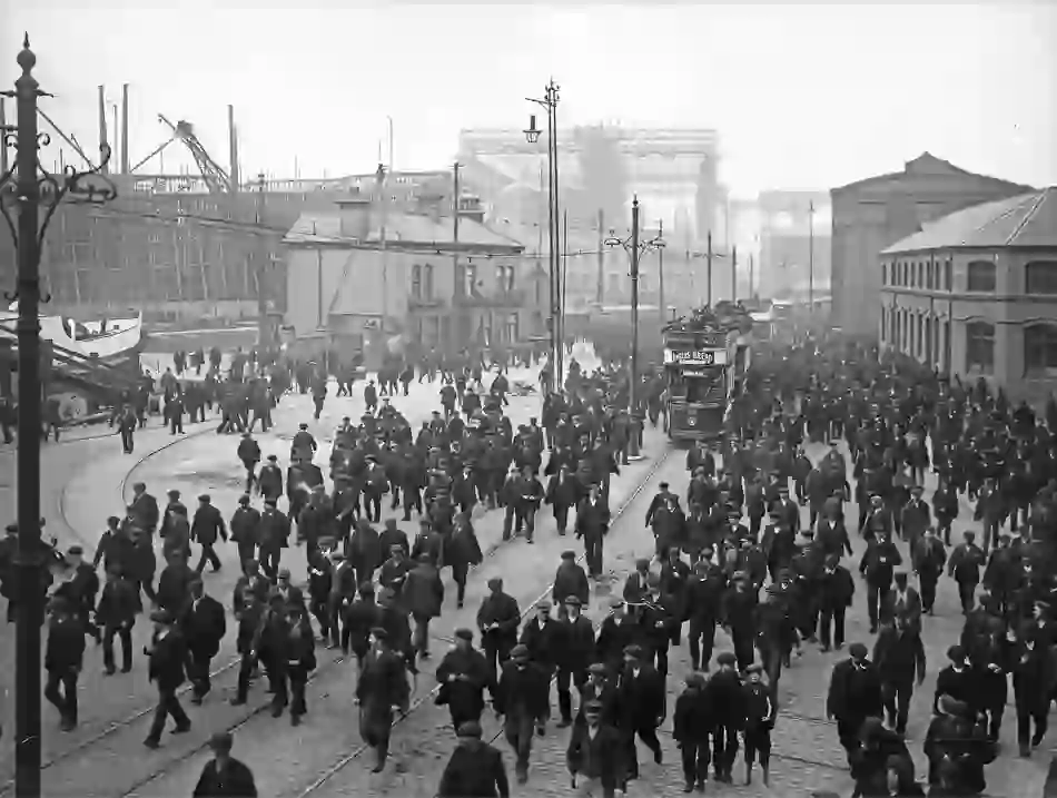 Crowds of shipyard workers leaving Harland &amp; Wolff, Titanic under construction visible behind them.