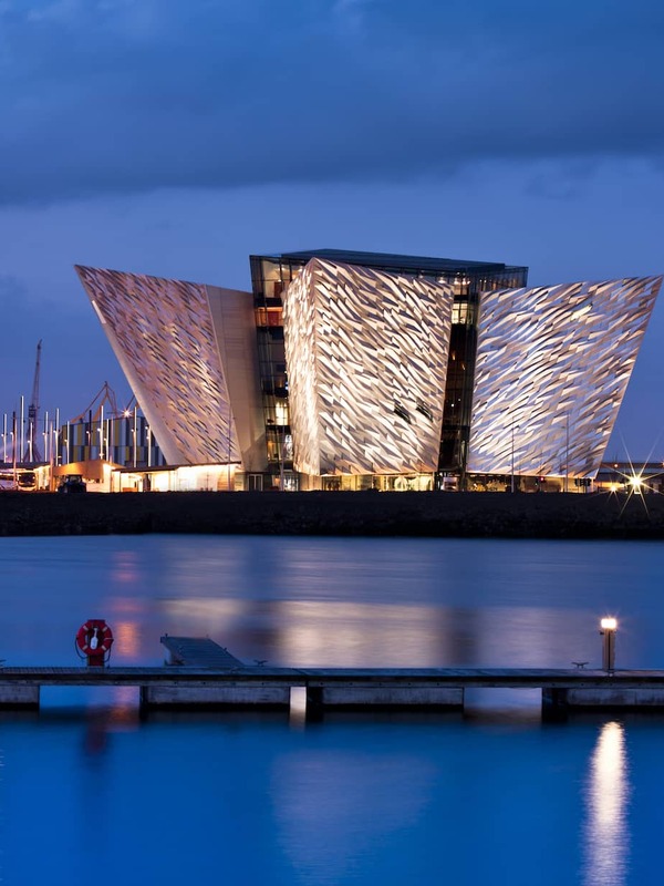 Titanic Belfast lit up at dusk, its angular facade reflected in the water, with a dark blue sky behind.