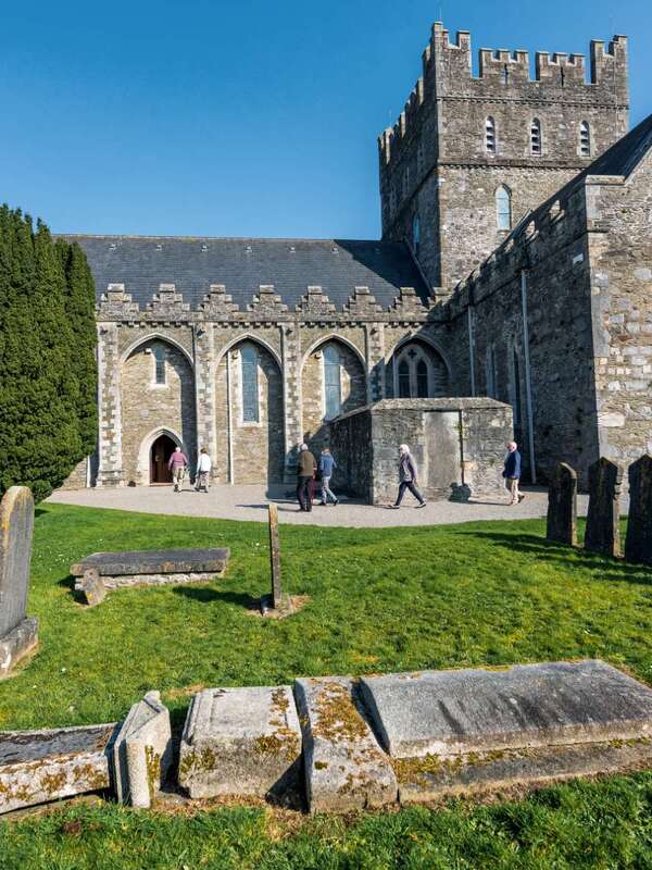 Visitors walking outside St Brigid’s Cathedral in Kildare, with round tower and gravestones under a clear blue sky.