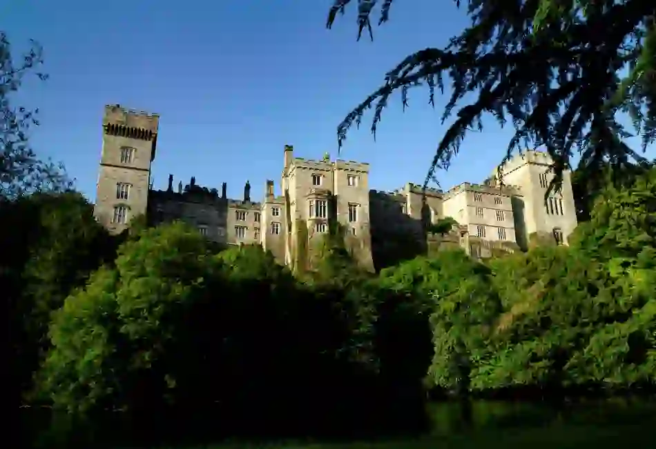 Lismore Castle with battlements rising above treetops and riverbank under a clear blue sky in Waterford.