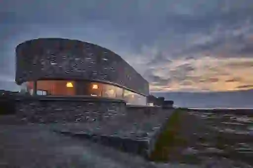 Low curved Inis Meáin Restaurant & Suites with stone cladding and panoramic windows on a rocky coast at sunset in Galway.