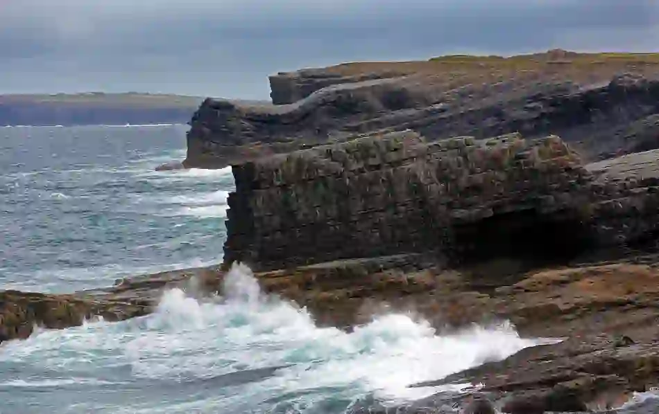 Waves crash against the folded sedimentary rock formations at Loop Head in County Clare under a grey sky.