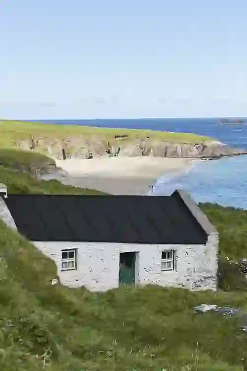 Traditional stone cottage with dark roof on Great Blasket Island, overlooking white sand beach and the Atlantic Ocean beyond.