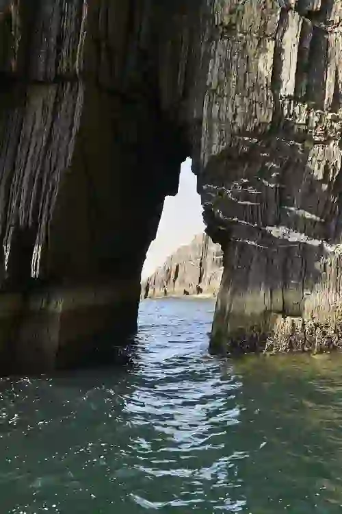 Narrow sea arch cut through layered black cliffs, revealing clear blue water and more jagged rock formations beyond.