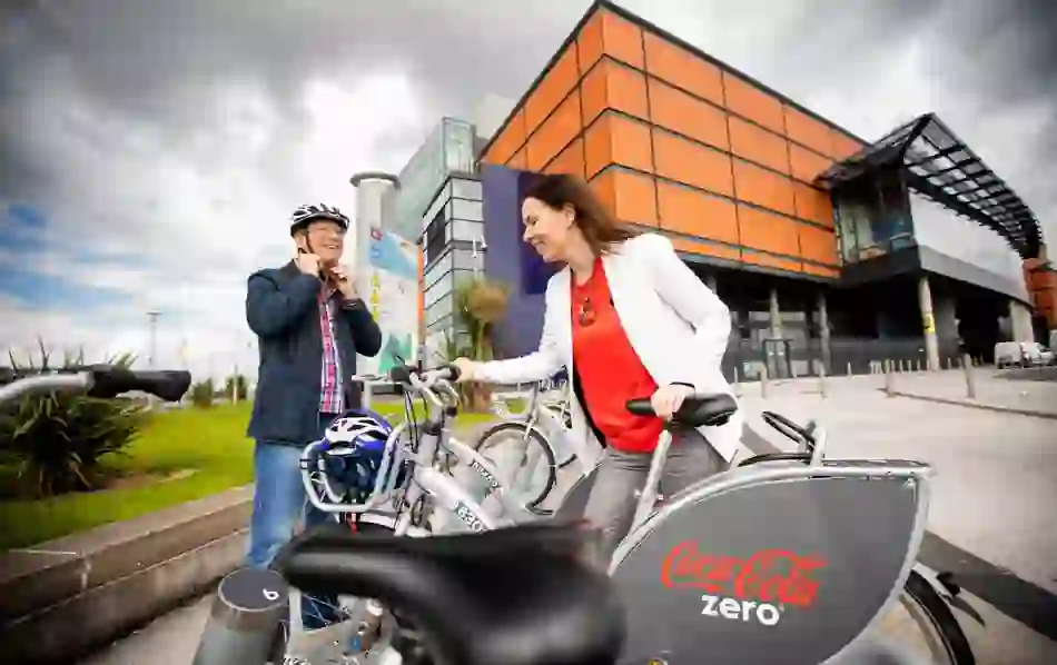 Two people prepare to cycle from a Belfast bike-share station outside the Odyssey Complex, promoting green urban travel.