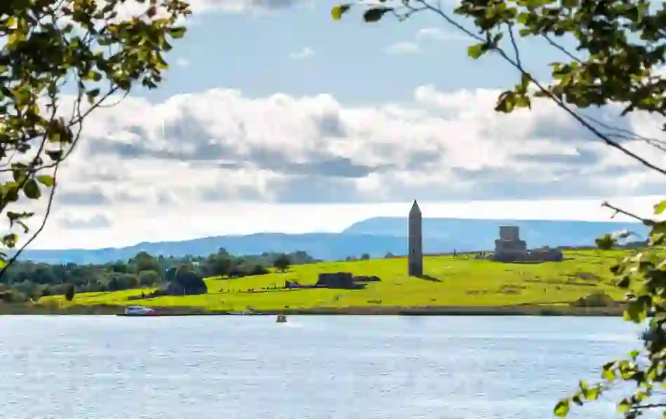 A round tower and abbey ruins rise from green fields on Devenish Island, framed by trees and seen across Lough Erne.