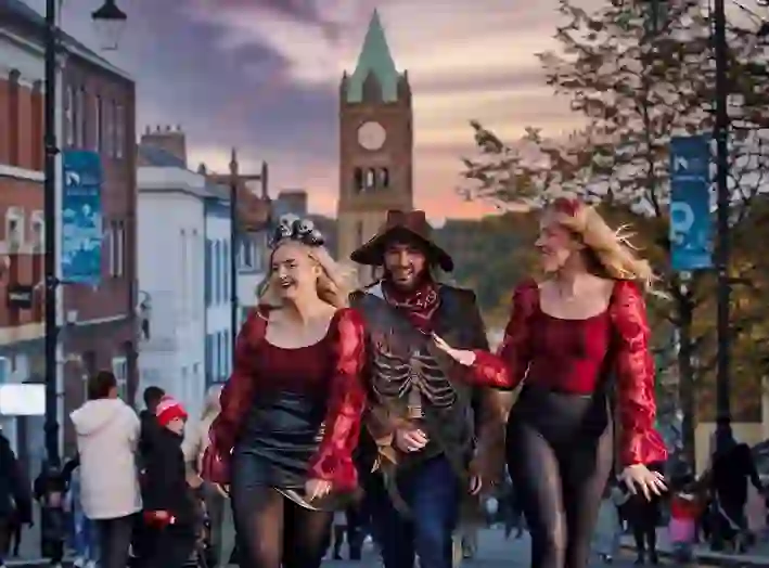 People in costume walk through the streets of Derry during Halloween celebrations, with the Guildhall clock behind.