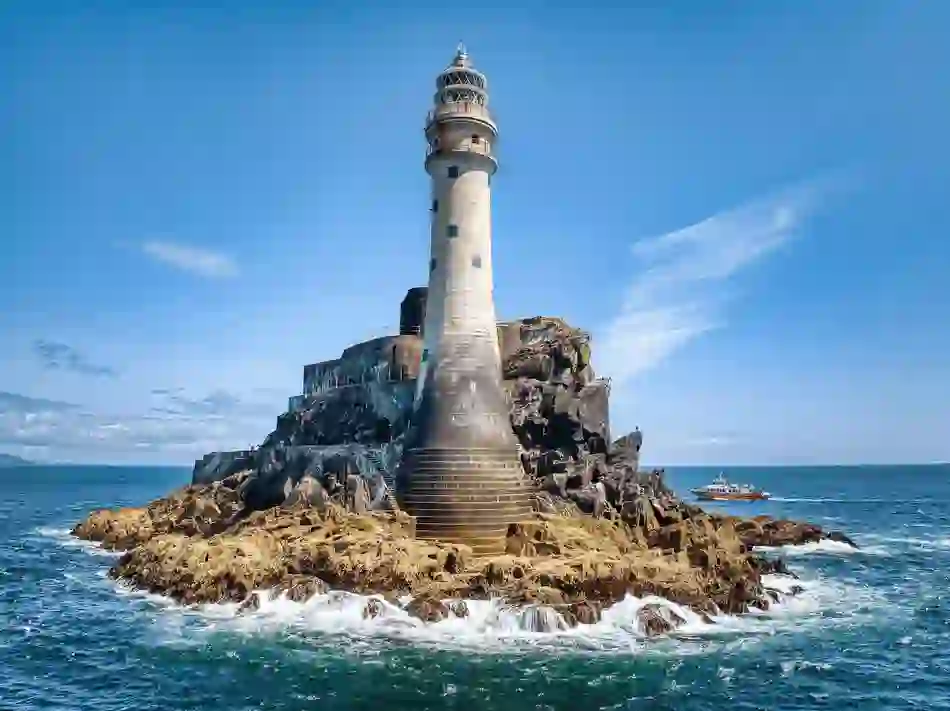 Fastnet Lighthouse rises from a rocky outcrop off the Cork coast, with waves crashing and a boat nearby.