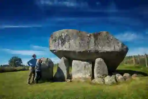 Two people standing beside Brownshill Dolmen in a grassy field under a deep blue sky in Carlow.