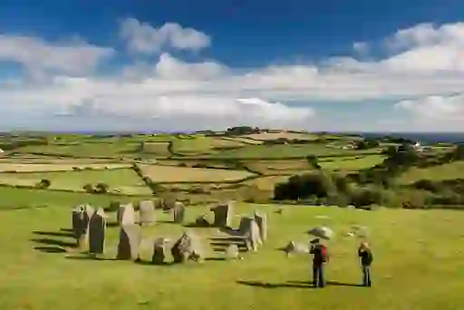 Two people looking at Drombeg stone circle on a grassy slope above patchwork farmland and sea in Cork.