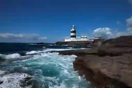 Hook Lighthouse on the Wexford coast with waves crashing against the rocky shore under a blue sky.