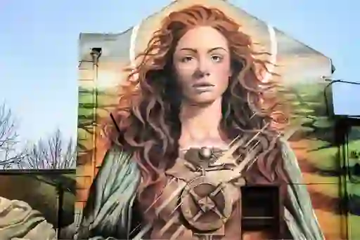  Large mural of Saint Brigid painted on a building wall, featuring symbolic elements and flowing red hair.
