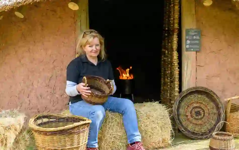 Woman weaving and displaying baskets outside a thatched building with a fire inside.