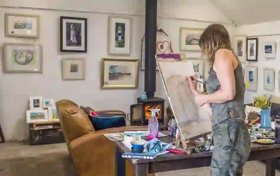 Artist painting at an easel in a cosy Irish studio with framed artwork on the walls.