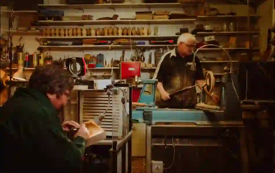 Two craftsmen working with wood in a workshop filled with tools and shelves.