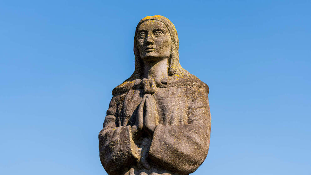 Stone statue of St Brigid with hands clasped in prayer against a clear blue sky in Kildare.