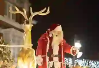 Santa in a red suit waving from a festive float beside a golden reindeer during a Christmas celebration.